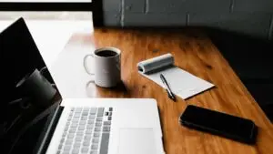 A small business owner writing blog post ideas on a notepad next to a laptop at a shop counter.