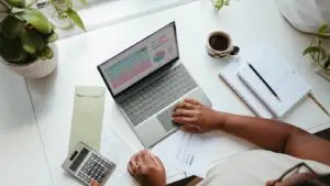 A small business owner and a marketing consultant review a monthly budget spreadsheet on a laptop at a shop counter.